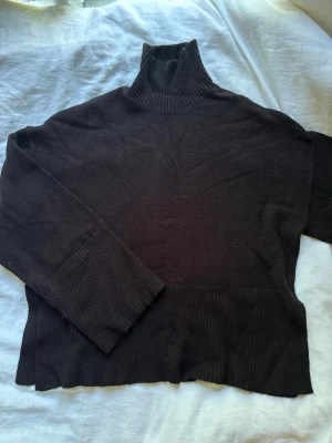 Svart polotröja i ribbstickat - Mysig svart stickad polotröja med bred ribbstickning vid hals, ärmslut och nederkant. Tröjan har lång ärm och en hög polokrage som ger en stilren look. Perfekt för kalla dagar och enkel att matcha med olika outfits.