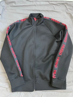 Svart ziptröja från Hugo Boss - Svart ziptröja från Hugo Boss med röd logga längs ärmarna och hög krage. Tröjan har hel dragkedja framtill och ribbade muddar vid ärmslut och nederkant. Perfekt för en sportig och trendig look.