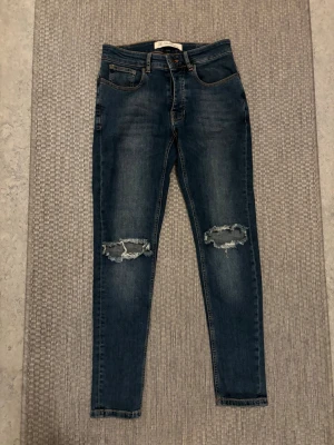 Blå slitna jeans från The Blue Uniform - Snygga blå jeans från The Blue Uniform med skinny fit och slitningar över knäna. Klassisk femficksmodell med orangea sömmar och normal midja. Perfekta för en avslappnad och trendig look.