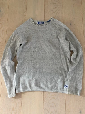 Ljusgrå stickad tröja Jack & Jones - Ljusgrå stickad tröja från Jack & Jones med rund halsringning och diskret struktur i materialet. Tröjan har långa ärmar och en liten logolapp nertill vid fållen. Perfekt för lager på lager och enkel att matcha.