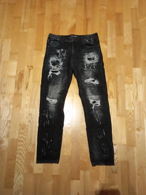 Svarta slitna skinny jeans (supply and demand) - Säljer ett par svarta skinny jeans med kraftiga slitningar och hål på framsidan samt vita färgstänk för en riktigt edgy look. Jeansen har fem fickor och normal midja. Perfekta för dig som gillar streetstyle och vill sticka ut.