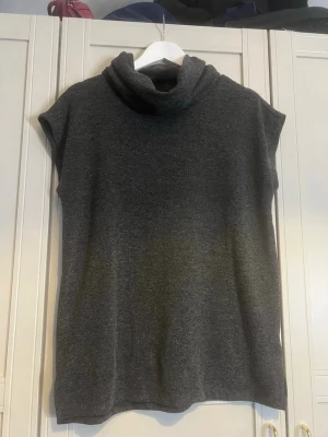 Mörkgrå stickad ärmlös polotröja - Mysig mörkgrå stickad ärmlös polotröja med lös passform och hög krage. Perfekt att styla över en långärmad topp eller bära som den är. Materialet känns mjukt och följsamt, och modellen har en enkel, stilren look.