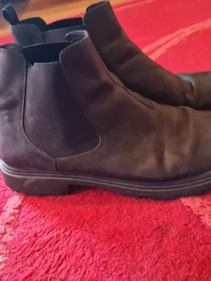 Svarta Chelsea boots i mocka - Säljer ett par klassiska svarta Chelsea boots i mocka med elastiska sidopaneler och låg klack. Skorna har rundad tå och diskret sula, perfekta för en stilren look. Passar till både jeans och kostym.