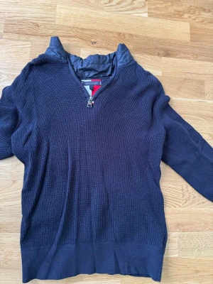 Mörkblå stickad tröja med halv dragkedja från Tommy hilfiger - Mörkblå stickad tröja med halv dragkedja och hög krage i unikt quiltat material. Tröjan har långa ärmar och ribbade muddar. Snygg detalj med quiltad panel över axlar och krage, perfekt för en cool och avslappnad stil.