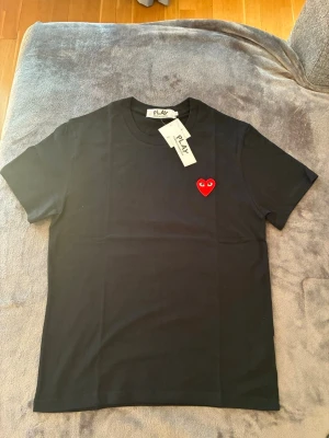 Comme des Garçons T-shirt  - Comme des Garçons T-shirt. Snygg svart t-shirt med korta ärmar och ett broderat rött hjärta med ögon på bröstet. Tillverkad i mjuk bomull och har en klassisk passform. Perfekt för dig som gillar stilrena plagg med en twist. 