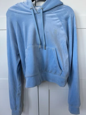 Ljusblå velour hoodie med strass - Säljer en ljusblå hoodie i mjuk velour med huva och dragskedja. Framtill finns en stor magficka och på ryggen står det 'Juicy' i glittriga strasstenar. Hoodien har långa ärmar och bred mudd nedtill, perfekt för en chill och trendig look. Säljs då den inte kommer till användning längre! 