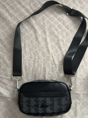 Svart axelväska från Lacoste - Snygg svart axelväska från Lacoste med justerbar bred rem och silverfärgade detaljer. Väskan har ett mönster med Lacoste-loggor på framsidan och dragkedja upptill. Perfekt för att bära det viktigaste med stil.