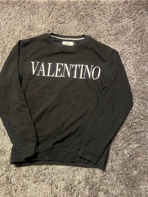 Svart sweatshirt från Valentino - Stilren svart sweatshirt från Valentino med stor vit logga framtill. Tröjan har rund halsringning, långa ärmar och ribbade muddar vid ärmslut och nederkant. Perfekt för en trendig och avslappnad look.
