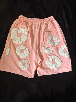 Denim tears shorts - Rosa denim tears shorts med elastisk midja. Dekorerade med stora vita pufftryck i cirkelformade mönster på både fram- och baksida. Tillverkade i mjukt bomullsliknande material och har en bakficka. Perfekta för en avslappnad och trendig stil.