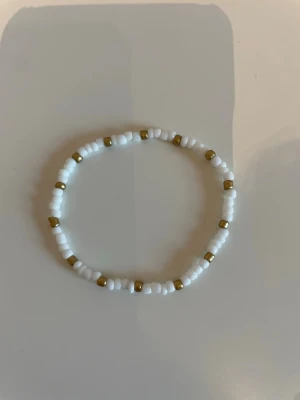 Vitt och guldigt pärlarmband - Säljer ett stilrent armband med vita och guldfärgade pärlor. Armbandet är elastiskt och har en enkel, rund design som passar till många olika stilar. Perfekt accessoar för dig som gillar minimalistiska smycken.