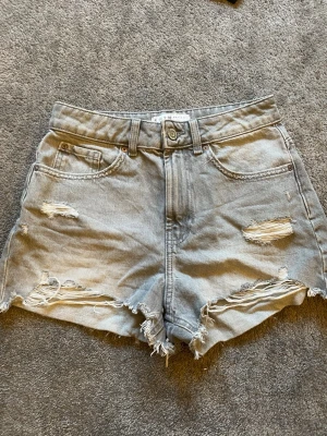 Gråa högmidjade jeansshorts - Säljer ett par gråa högmidjade jeansshorts med slitna detaljer och fransig kant. Shortsen har klassisk femficksdesign, bälteshällor och knappgylf. Perfekta för en avslappnad och trendig sommarlook.