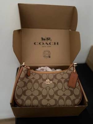 Coach brun teri shoulder bag signature canvas - Snygg brun handväska från Coach med klassiskt monogrammönster och detaljer i ljusbrunt läder. Väskan har guldfärgade metalldetaljer, avtagbar axelrem och Coach-tag. Perfekt storlek för det viktigaste och en stilren look.