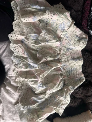 Pastellfärgad volangkjol med spets - Söt kortkjol i pastellfärger med volanger och broderad spetskant. Kjolen har ett smockat midjeparti, luftigt tyg med diskret blommigt mönster.