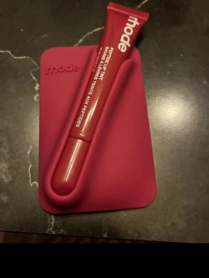 Rhode Peptide Lip Tint röd - Säljer ett Rhode Peptide Lip Tint i en snygg röd nyans. Läppbalsamet kommer i en tub med vit text och har en modern, stilren design. Perfekt för dig som vill ha återfuktade läppar med en hint av färg.