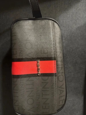 Svart necessär med röd detalj Valentino - Snygg svart necessär från Valentino med diskret logomönster och en bred röd rand framtill. Väskan har en rektangulär form och är tillverkad i ett tåligt konstlädermaterial. Perfekt för att hålla ordning på småsaker eller som smidig reseväska.
