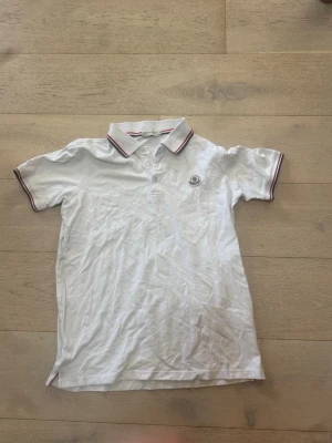 Vit pikétröja från Moncler - Klassisk vit pikétröja från Moncler med korta ärmar och krage med röda och blå ränder. Tröjan har knappslå framtill och Moncler-logga på bröstet. Perfekt till en clean och sportig stil.