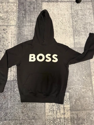 Svart hoodie med BOSS-tryck - Säljer en svart hoodie med stor vit BOSS-logga på bröstet. Klassisk modell med huva och känguruficka framtill. Perfekt för dig som vill ha en enkel men snygg streetwear-look.
