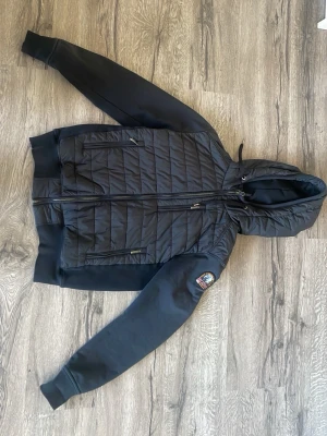 Svart parajumpers pufferjacka med huva - Svart pufferjacka från parajumpers  med quiltad framsida och huva. Jackan har dragkedja, två fickor fram och en patch på ena ärmen. Ärmar och rygg är i mjukare material, vilket ger en sportig look.