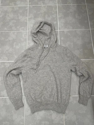 Kashmir hoodie - Mysig grå stickad hoodie med huva och dragsko. Tröjan har långa ärmar, ribbade muddar vid ärmslut och nederkant. Perfekt för dig som gillar en enkel och stilren look. Materialet ser ut att vara mjukt och värmande, perfekt för kyliga dagar. Har krympt i tvätten så passa typ xs