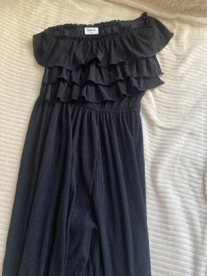 Svart off shoulder jumpsuit Kappahl - Supersnygg svart jumpsuit från Kappahl i storlek 146. Off shoulder-modell med flera volanglager upptill och vida ben. Tillverkad i ett mjukt, plisserat tyg som ger en skön och luftig känsla. Perfekt för dig som vill ha en trendig och bekväm look.