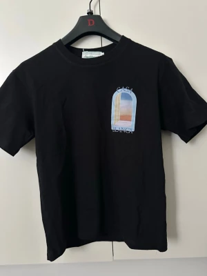 Svart t-shirt med tryck från Casablanca - Snygg svart t-shirt från Casablanca med ett färgglatt tryck på bröstet som föreställer ett fönster med solnedgång. Klassisk passform och rund halsringning. Perfekt för dig som vill ha en stilren men ändå unik look.