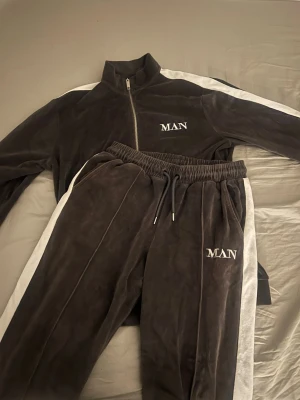 Mörkgrå velour loungewear-set MAN - Säljer ett mörkgrått loungewear-set i mjuk velour med vita ränder längs ärmar och ben. Setet består av ziptröja och byxor med resår i midjan och dragsko. Båda delarna har broderad texten 'MAN' på bröstet och benet. Perfekt för chill dagar hemma.