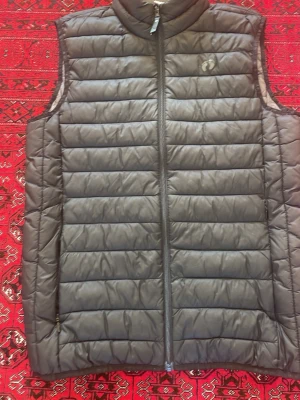 Grå dunväst från Hangten - Snygg grå dunväst från Hangten med quiltad design och hög krage. Västen har dragkedja framtill och två sidofickor med dragkedja. Perfekt att slänga över en hoodie eller tröja för extra värme. Tillverkad i lätt och mjukt material.