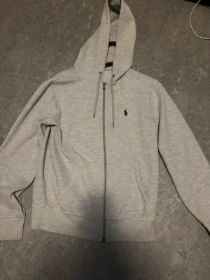 Ljusgrå hoodie från Polo Ralph Lauren - Snygg ljusgrå hoodie från Polo Ralph Lauren med dragkedja framtill, två fickor och klassisk huva med dragsko. Ikoniska lilla loggan broderad på bröstet. Tillverkad i mjukt bomullsmaterial som är skönt att ha på sig. Perfekt för en avslappnad och stilren look.