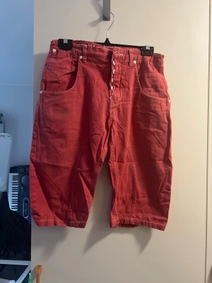 Röda loose shorts med knappar - Snygga röda shorts i loose fit med flera framfickor och knappgylf. Shortsen har bälteshällor och är tillverkade i ett slitstarkt bomullstyg. Perfekta för sommardagar och ger en avslappnad vibe. Saknar en knapp fram!