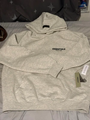 Ljusgrå Essentials Fear of God hoodie - Säljer en ljusgrå hoodie från Essentials Fear of God med svart logga på bröstet. Hoodien har huva, ribbade muddar och en stor magficka. Tillverkad i mjukt bomullsmaterial och har en clean, oversized look som passar perfekt till streetwear.