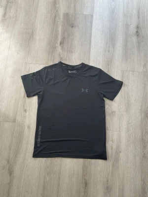 Svart tränings-tshirt från Under Armour - Snygg svart t-shirt från Under Armour med diskret logga på bröstet och tryck på axeln. Tillverkad i ett lätt och snabbtorkande syntetmaterial som passar perfekt till träning. Klassisk passform med korta ärmar och rund hals.