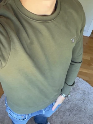 Olivgrön sweatshirt från GANT - Säljer en olivgrön sweatshirt från GANT med rund halsringning och broderad logga på bröstet. Tröjan har ribbade muddar vid ärmslut och nederkant samt klassisk passform. Perfekt för en avslappnad och stilren look.