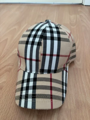 Beige keps med rutigt mönster - Snygg keps i beige med klassiskt rutigt mönster i svart, vitt och rött. Kepsen har böjd skärm och justerbar baksida. Perfekt accessoar för en trendig streetstyle-look.