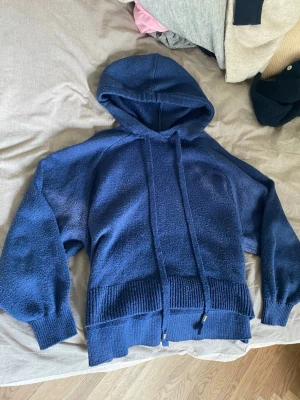 Mörkblå stickad hoodie - Mysig mörkblå stickad hoodie med huva och långa snören. Tröjan har breda ribbade muddar vid ärmslut och nederkant, samt en avslappnad passform. Perfekt för chill dagar eller när du vill vara extra bekväm.