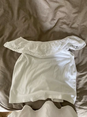 Vit offshoulder topp med spets - Söt vit offshoulder topp med bred volangkant i spets och broderade detaljer. Toppen är ärmlös och har en luftig passform, perfekt för varma dagar. Volangen ger en romantisk vibe och toppen är lätt att matcha till olika outfits.