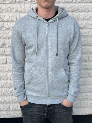 Ljusgrå zip hoodie med huva - Säljer en ljusgrå hoodie med dragkedja och huva. Hoodien har två fickor framtill, justerbara snören i huvan och ribbade muddar vid ärmar och midja. Tillverkad i mjukt sweatshirtmaterial som är riktigt skönt att ha på sig. Storlek XS, skulle nog även passa S. 