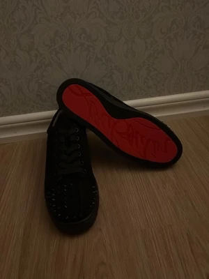 Svarta sneakers med nitar och röd sula - Säljer ett par svarta sneakers med coola nitar på tån och ikonisk röd sula. Skorna har snörning och en stilren, låg profil som passar till streetwear och trendiga outfits. Materialet ser ut att vara läder eller liknande för en lyxig känsla.