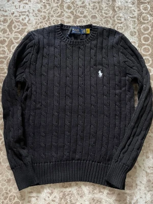 Svart kabelstickad tröja Polo Ralph Lauren - Kabelstickad svart tröja från Polo Ralph Lauren med rund halsringning och vit broderad logga på bröstet. Tröjan har ribbade muddar vid ärmslut och nederkant, perfekt för dig som gillar klassisk och stilren design. Tröjan är i storlek M men passar även som S