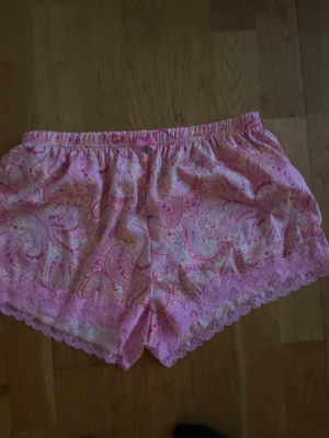 Rosa pyjamasshorts med spetsdetaljer - Säljer ett par söta pyjamasshorts i ljusrosa med paisleymönster och dekorativa spetskanter. Shortsen har resår i midjan och en liten rosett framtill. Perfekta för mysiga hemmakvällar.