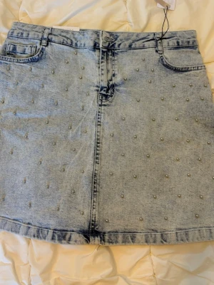 Ljusblå jeanskjol med pärlor New Look - Trendig ljusblå jeanskjol från New Look med pärldetaljer över hela kjolen. Klassisk femficksmodell och kort passform. Perfekt för dig som vill ha en cool och unik denimlook.