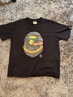 Svart Bape t-shirt med tryck - Bape / A Bathing Ape "Ape Head Chain" T-shirt (M) . Svart t-shirt från Bape med stort grafiskt tryck framtill som visar en figur med gul keps, cigarr och guldkedja. Klassisk passform och rund halsringning. Perfekt för dig som gillar streetwear och vill sticka ut.