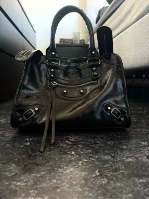 Svart handväska i skinn med detaljer - Snygg svart handväska i skinn med silverfärgade metalldetaljer, dragkedja framtill och dekorativa spännen. Väskan har dubbla handtag, varav ett är flätat, och långa läderband som dekoration. Perfekt för dig som gillar en edgy och stilren look.