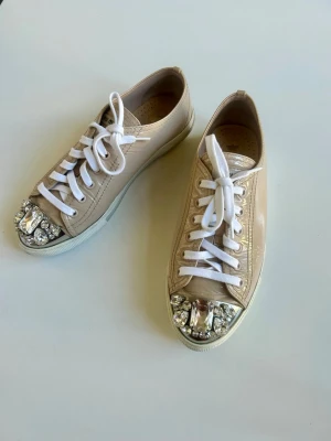 Miu Miu Crystal Cap-Toe Sneakers - Säljer ett par fantastiska och exklusiva sneakers från det italienska lyxmärket Miu Miu. Modellen är tillverkad i ett elegant ljusbeige lackskinn (patent leather)   Från Miu Mius ikoniska kollektion med gnistrande kristall- och strassdetaljer framme på tåkappan.  Märke: Miu Miu Modell: Crystal Cap-Toe Sneakers Färg: Nude / Ljusbeige  Material: Lackskinn (Patent leather) med tåkappa i metall och kristaller Storlek: 38 Skick: Mycket bra skick, endast sparsamt använda. Alla kristaller är intakta.