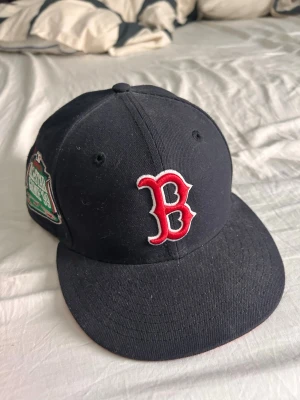 Boston Red Sox svart keps - Snygg svart keps från New Era med Boston Red Sox-logga i rött och vitt framtill. Kepsen har röd undersida på skärmen och broderad patch på sidan. Modellen är 59FIFTY och materialet är bomull. Perfekt för dig som gillar sportig stil.