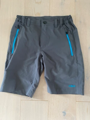 Grå shorts med blå detaljer från CMP - Snygga grå shorts från CMP med blå dragkedjor på fickorna och logga framtill. Shortsen har normal passform, bälteshällor och elastisk midja för extra komfort. Perfekta för sommarens alla äventyr.