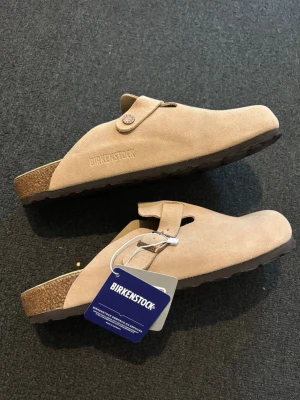 Birkenstock tofflor - Säljer ett par helt nya beiga tofflor från Birkenstock i mjuk mocka. De har en klassisk slip-in modell med justerbar rem och korksula för extra komfort. Perfekta för dig som vill ha både stil och bekvämlighet i vardagen.