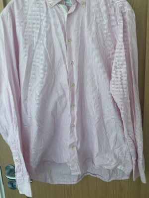 Rosa och vit randig skjorta - Säljer en klassisk långärmad skjorta med tunna rosa och vita ränder. Skjortan har knappar hela vägen framtill och en avslappnad passform. Perfekt till jeans eller chinos för en clean look.