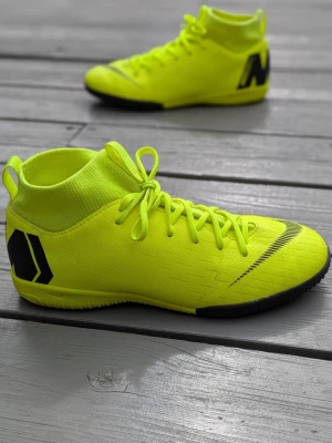 Neongula Nike fotbollsskor - Säljer ett par neongula Nike fotbollsskor med svart sula och svarta detaljer. Skorna har högt skaft i strumpliknande material och snörning för bra passform. Perfekta för dig som vill sticka ut på planen och ha grym kontroll på bollen.