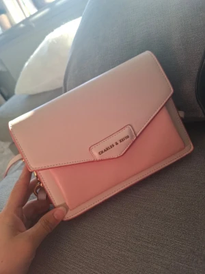 Ljusrosa kuvertväska Charles & Keith - Snygg ljusrosa kuvertväska från Charles & Keith i slätt konstläder. Väskan har ett stilrent kuvertlock med magnetknäppning och flera fack inuti, inklusive ett med dragkedja. Perfekt för att bära det viktigaste och ger en fräsch touch till din outfit.