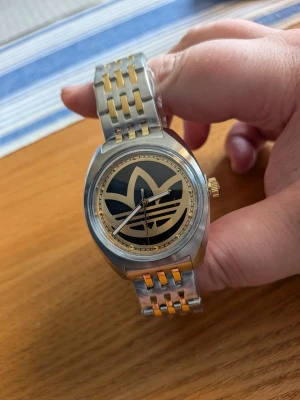 Adidas klocka i silver och guld - Stilren klocka från Adidas med metallarmband i silver och guldton. Urtavlan är svart med Adidas logga i guld, och boetten är rund i rostfritt stål. Klockan har guldfärgad krona och detaljer, perfekt för dig som gillar sportig och trendig stil.
